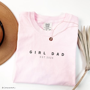 May include: A light pink t-shirt with the words "GIRL DAD EST. 2026" printed in black. A gold necklace with a circular pendant rests on the shirt. A brown hat and a dried plant are in the background.