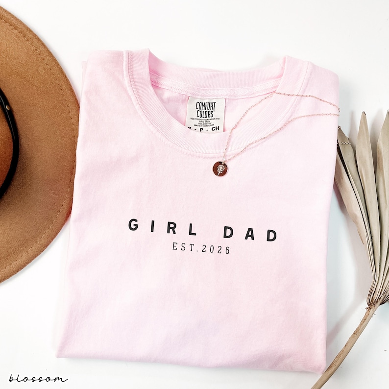 May include: A light pink t-shirt with the words "GIRL DAD EST. 2026" printed in black. A gold necklace with a circular pendant rests on the shirt. A brown hat and a dried plant are in the background.