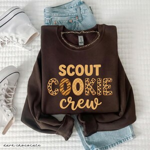 May include: A dark chocolate-colored sweatshirt with the words "SCOUT COOKIE crew" in a tan font. The word "COOKIE" has a leopard print design. The sweatshirt is paired with light blue jeans and white sneakers.