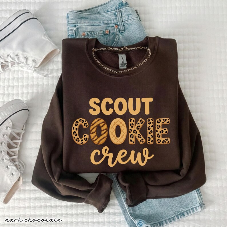 May include: A dark chocolate-colored sweatshirt with the words "SCOUT COOKIE crew" in a tan font. The word "COOKIE" has a leopard print design. The sweatshirt is paired with light blue jeans and white sneakers.