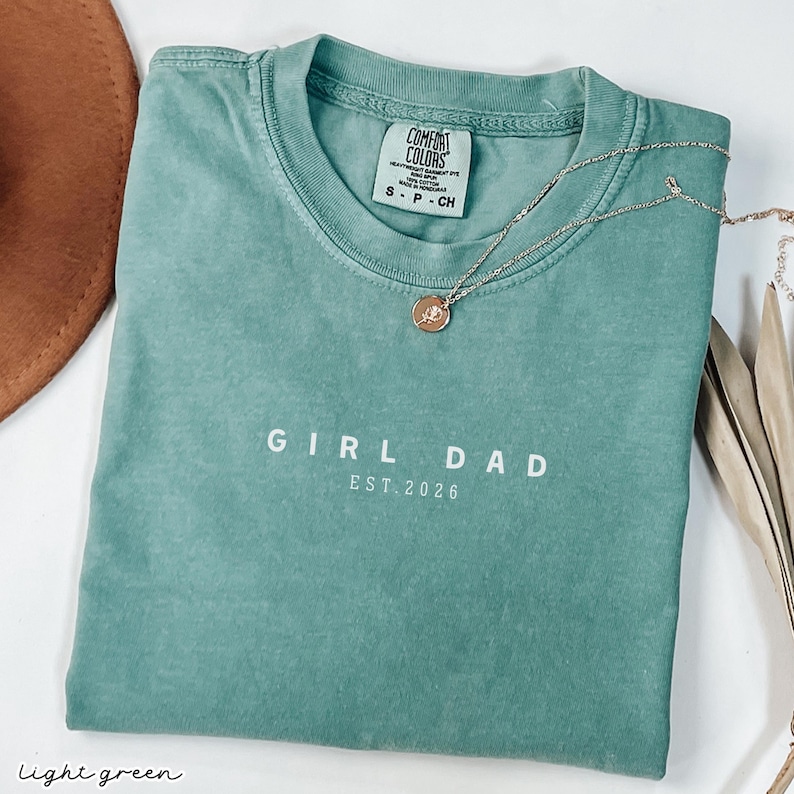 May include: A light green t-shirt with the words "GIRL DAD EST. 2026" printed in white. A gold necklace with a pendant is draped across the shirt. The shirt has a Comfort Colors tag.