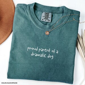 May include: A teal blue t-shirt with the text "proud parent of a dramatic dog" in white lettering. The shirt is made of a soft, comfortable material and has a relaxed fit.