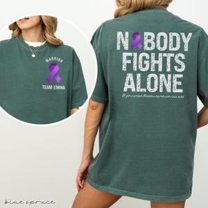 Pode incluir: Uma t-shirt verde-azulado com o texto "NOBODY FIGHTS ALONE" e uma fita roxa nas costas. A frente da t-shirt tem uma fita roxa e as palavras "WARRIOR" e "TEAM EMMA". A hashtag #pancreaticancerawareness tamb&eacute;m &eacute; vis&iacute;vel.