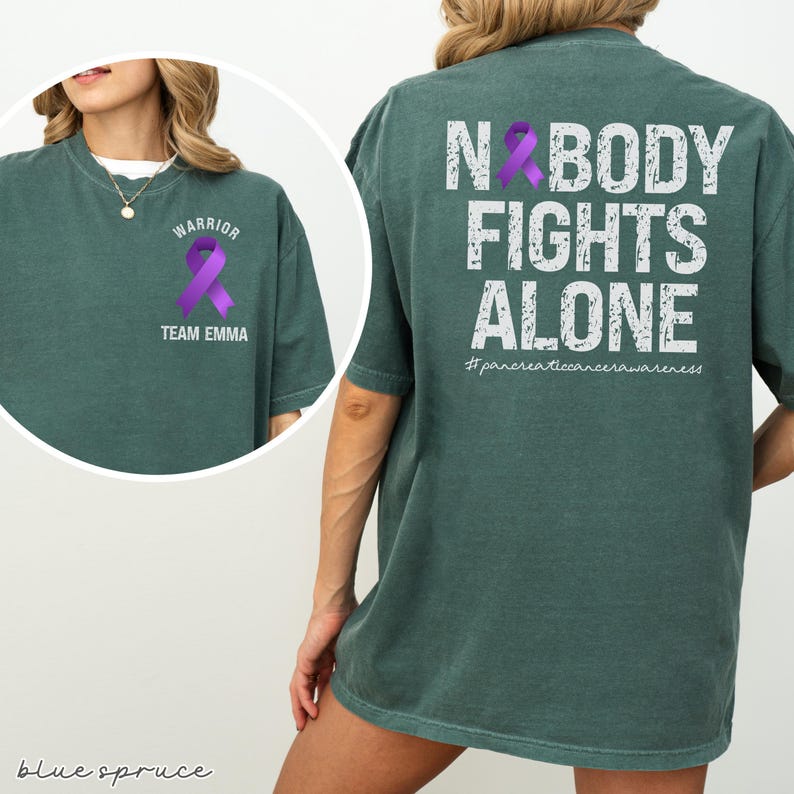 Pode incluir: Uma t-shirt verde-azulado com o texto "NOBODY FIGHTS ALONE" e uma fita roxa nas costas. A frente da t-shirt tem uma fita roxa e as palavras "WARRIOR" e "TEAM EMMA". A hashtag #pancreaticancerawareness tamb&eacute;m &eacute; vis&iacute;vel.