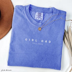 May include: A blue Comfort Colors t-shirt with the text "GIRL DAD EST. 2026" in white. A gold necklace with a round pendant rests on the shirt. A brown hat is partially visible in the background.