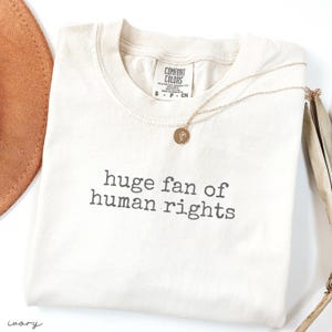 May include: Ivory-colored t-shirt with the phrase "huge fan of human rights" printed in gray. A gold necklace with a circular pendant rests on the shirt. The shirt is folded and has a Comfort Colors tag.