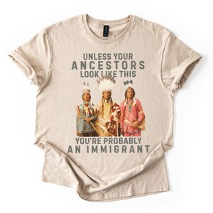 May include: Beige t-shirt with the text "UNLESS YOUR ANCESTORS LOOK LIKE THIS YOU'RE PROBABLY AN IMMIGRANT" above a graphic of three people in traditional Native American clothing. The shirt is folded.