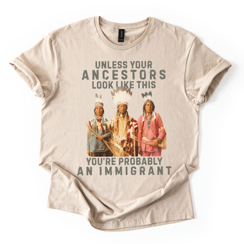 May include: Beige t-shirt with the text "UNLESS YOUR ANCESTORS LOOK LIKE THIS YOU'RE PROBABLY AN IMMIGRANT" above a graphic of three people in traditional Native American clothing. The shirt is folded.