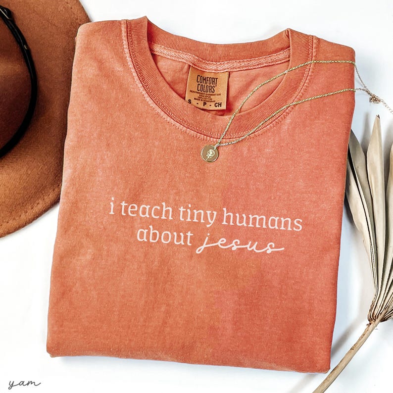 May include: A coral-colored t-shirt with the phrase "i teach tiny humans about jesus" in white script. A gold necklace with a pendant rests on the shirt. The shirt has a Comfort Colors tag.