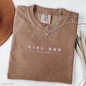 May include: Espresso-colored t-shirt with the words "GIRL DAD EST. 2026" printed in white. A silver necklace with a small pendant rests on the shirt. The shirt is folded and has a tag that says "Comfort Colors".