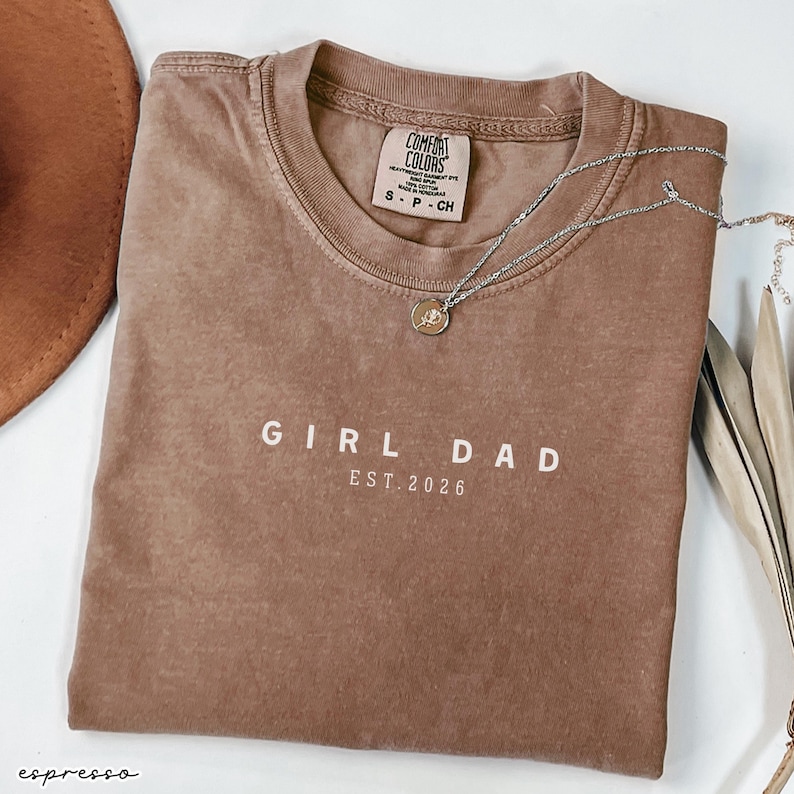 May include: Espresso-colored t-shirt with the words "GIRL DAD EST. 2026" printed in white. A silver necklace with a small pendant rests on the shirt. The shirt is folded and has a tag that says "Comfort Colors".