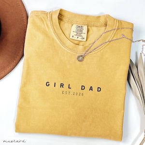 May include: Mustard-colored t-shirt with the text "GIRL DAD EST. 2026" in black. The shirt has a crew neck and short sleeves. A silver necklace with a small pendant is draped across the shirt. The shirt is labeled "Comfort Colors".