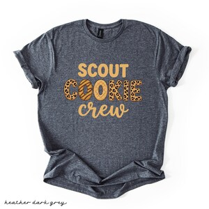 May include: Heather dark grey t-shirt with the words "SCOUT COOKIE crew" in a playful font. The word "SCOUT" is in a solid font, while "COOKIE" has a cookie-themed design, and "crew" is in a cursive font.