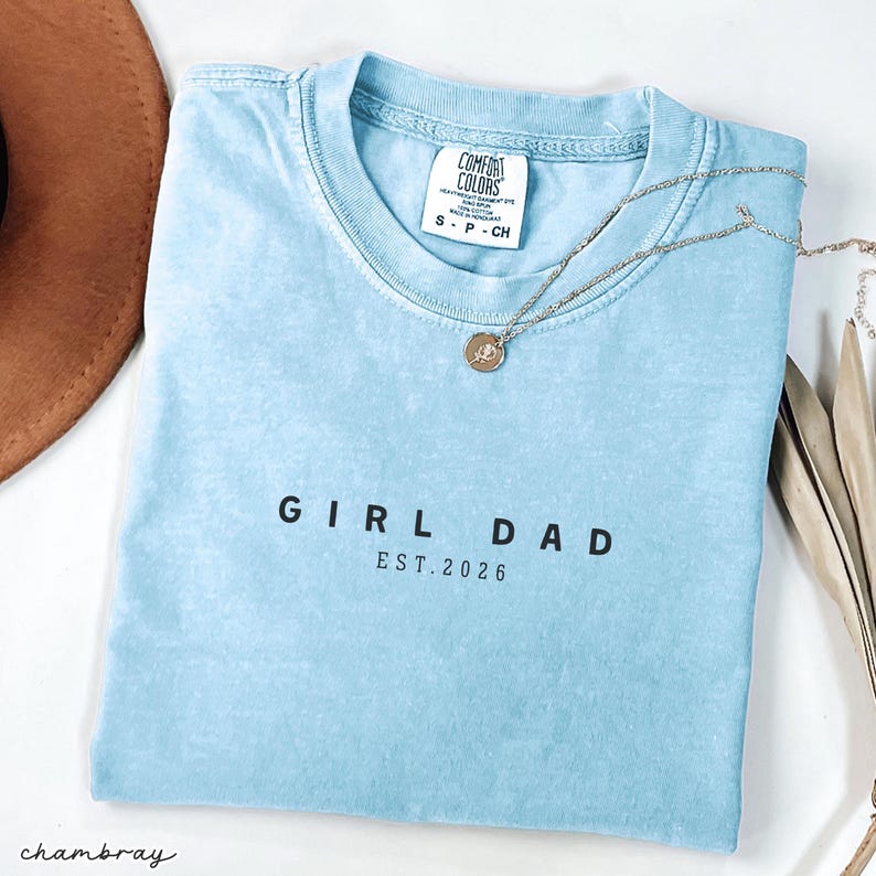 May include: A light blue t-shirt with the text "GIRL DAD EST. 2026" in black. A silver necklace with a small pendant rests on the shirt. The shirt is folded and placed on a white surface, with a brown hat visible.