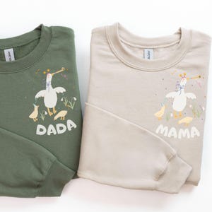 May include: Two sweatshirts, one olive green and one beige, are displayed. The green sweatshirt has the word "DADA" and a goose graphic. The beige sweatshirt has the word "MAMA" and a similar goose graphic. Both sweatshirts are crew neck style.