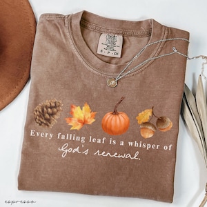May include: Brown Comfort Colors t-shirt with a fall-themed design. The shirt features a pinecone, maple leaf, pumpkin, and acorns. Text reads "Every falling leaf is a whisper of God's renewal."