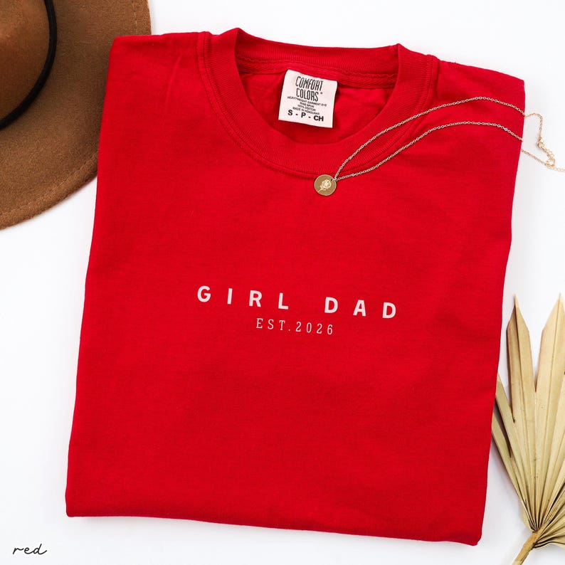 May include: A bright red sweatshirt with the words "GIRL DAD EST. 2026" printed in white. A gold necklace with a circular pendant rests on the shirt. The shirt has a Comfort Colors tag.
