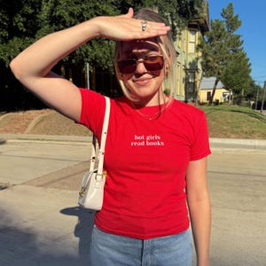 May include: A red t-shirt with the text "hot girls read books" in white. The shirt is paired with blue jeans and a white purse with a shoulder strap. The person is wearing sunglasses.