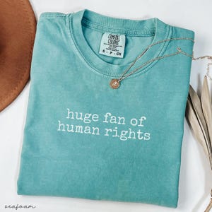 May include: Seafoam green t-shirt with the text "huge fan of human rights" in white. A gold necklace with a pendant rests on the shirt. The shirt is folded and has a tag at the neckline. A brown hat and dried leaves are in the background.