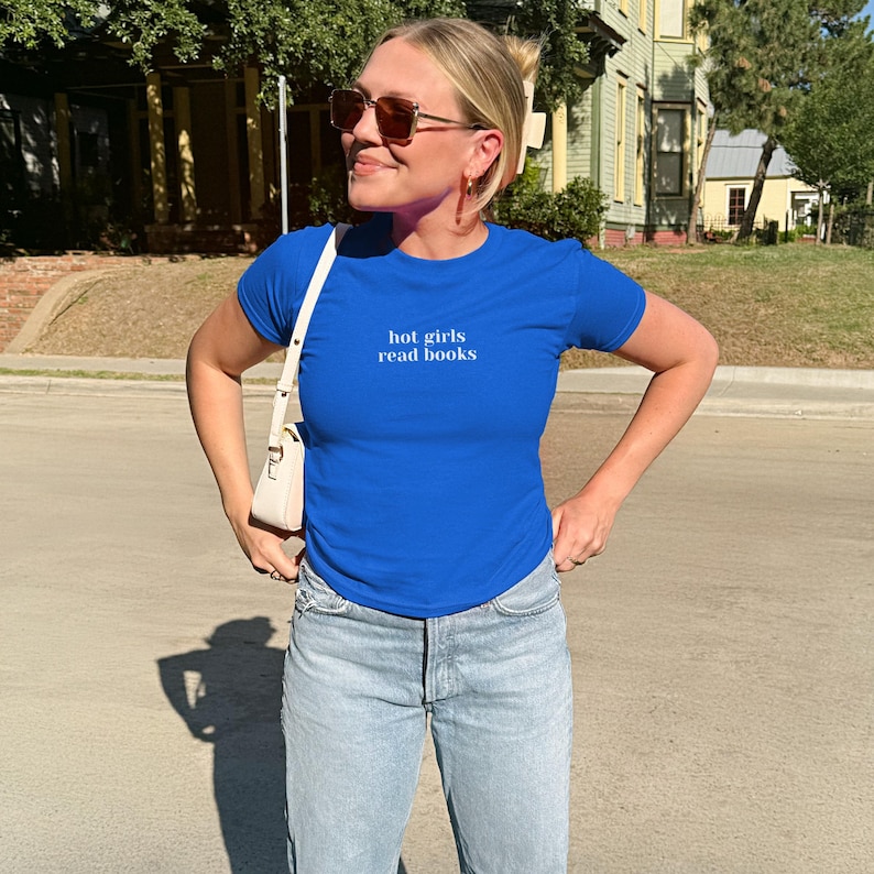 May include: A royal blue t-shirt with the text "hot girls read books" in white. The shirt is paired with light wash blue jeans and a white shoulder bag. The person is wearing sunglasses and has blonde hair.