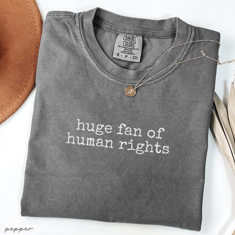 May include: A gray Comfort Colors t-shirt with the text "huge fan of human rights" in white. A silver necklace with a small pendant rests on the shirt. A brown hat is partially visible in the upper left corner.