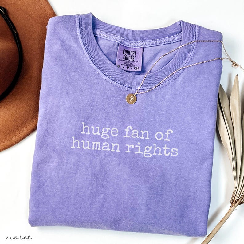May include: A violet Comfort Colors t-shirt with the text "huge fan of human rights" in white. A gold necklace with a pendant is draped across the shirt. A brown hat is visible in the background.