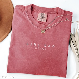 May include: Crimson-colored t-shirt with the text "GIRL DAD EST. 2026" in white. A silver necklace with a small pendant rests on the shirt. The shirt has a tag that reads "COMFORT COLORS". A brown hat is partially visible.