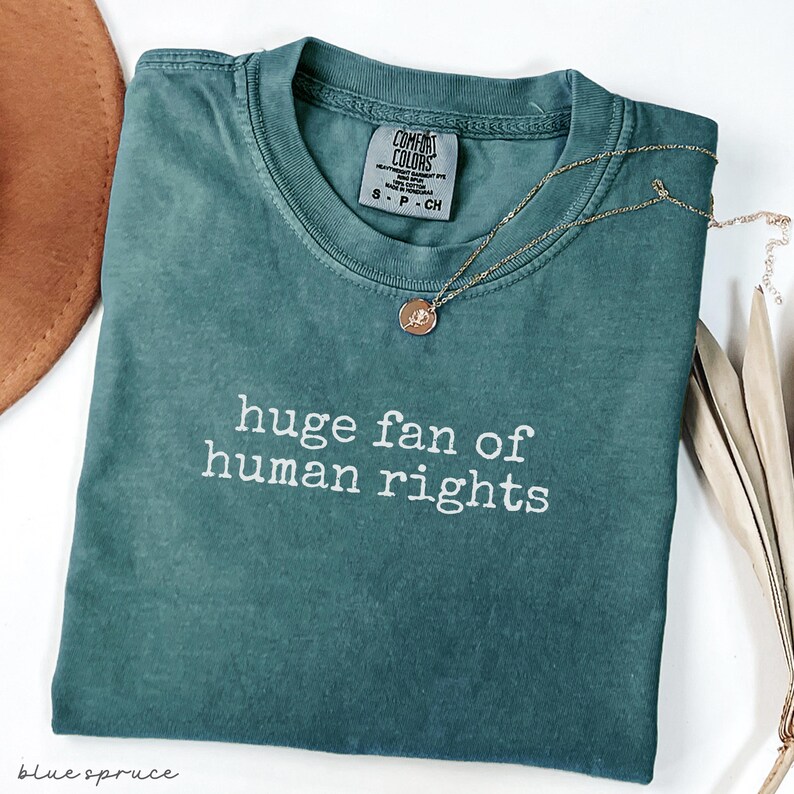 May include: A teal Comfort Colors t-shirt with the phrase "huge fan of human rights" in white lettering. A gold necklace with a pendant rests on the shirt. A brown hat and dried leaves are in the background.