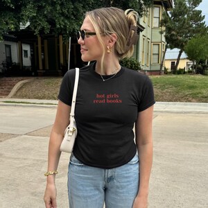 May include: Black crew neck t-shirt with the text "hot girls read books" in red. The shirt is paired with light blue jeans. The person is wearing sunglasses, gold earrings, and a gold bracelet. A white purse is over the shoulder.