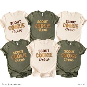May include: Six short-sleeved t-shirts in sand and heather olive colors. Each shirt features the text "SCOUT COOKIE crew" in a brown and tan design. The shirts are displayed on a white background.