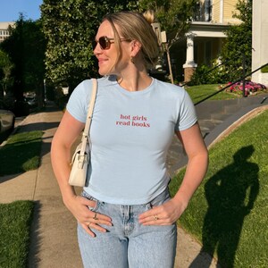 May include: Light blue t-shirt with the text "hot girls read books" in red. The shirt is paired with light wash blue jeans. A white purse is worn over the shoulder. The person is wearing sunglasses and rings.
