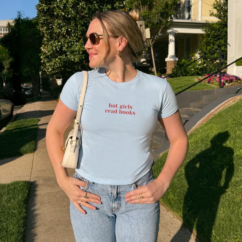 May include: Light blue t-shirt with the text "hot girls read books" in red. The shirt is paired with light wash blue jeans. A white purse is worn over the shoulder. The person is wearing sunglasses and rings.