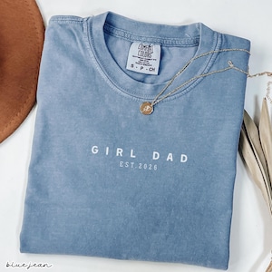 May include: A blue jean colored t-shirt with the words "GIRL DAD EST. 2026" printed in white. A gold necklace with a pendant rests on the shirt. The shirt is folded and has a Comfort Colors tag.