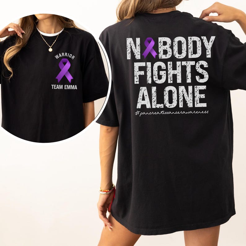 Pode incluir: Camiseta preta com uma fita de conscientiza&ccedil;&atilde;o roxa. A frente da camiseta diz "WARRIOR" acima da fita e "TEAM EMMA" abaixo. A parte de tr&aacute;s da camiseta diz "NOBODY FIGHTS ALONE" com uma fita roxa e a hashtag #pancreaticancerawareness.