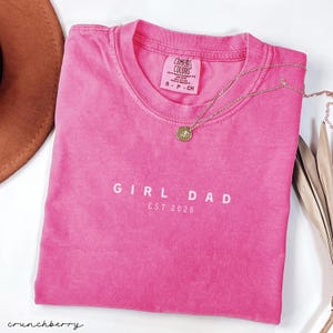 May include: A hot pink t-shirt with the words "GIRL DAD EST. 2026" printed in white. A gold necklace with a round pendant rests on the shirt. The shirt is folded and next to a brown hat.