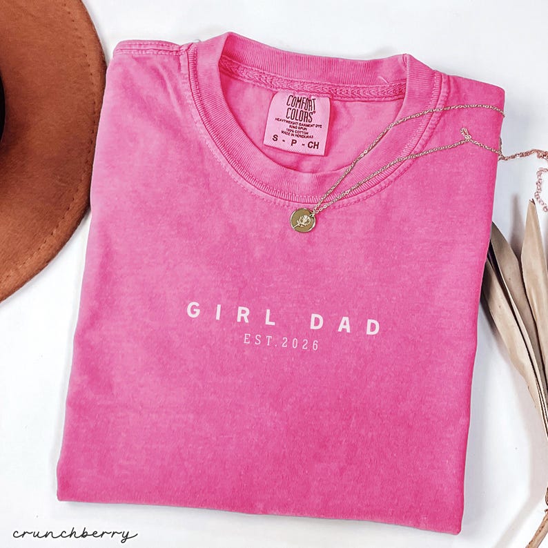 May include: A hot pink t-shirt with the words "GIRL DAD EST. 2026" printed in white. A gold necklace with a round pendant rests on the shirt. The shirt is folded and next to a brown hat.