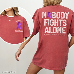 Pode incluir: Camiseta carmesim com a frase "NOBODY FIGHTS ALONE" e um la&ccedil;o roxo. A frente da camiseta tem um la&ccedil;o roxo e as palavras "WARRIOR" e "TEAM EMMA". A camiseta &eacute; de estilo casual.