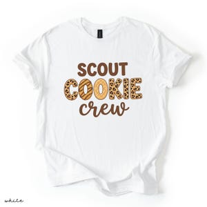 May include: White t-shirt with the words "SCOUT COOKIE crew" in brown lettering. The word "COOKIE" has a leopard print pattern and a cookie design. The shirt is made of a soft material and is perfect for cookie lovers.