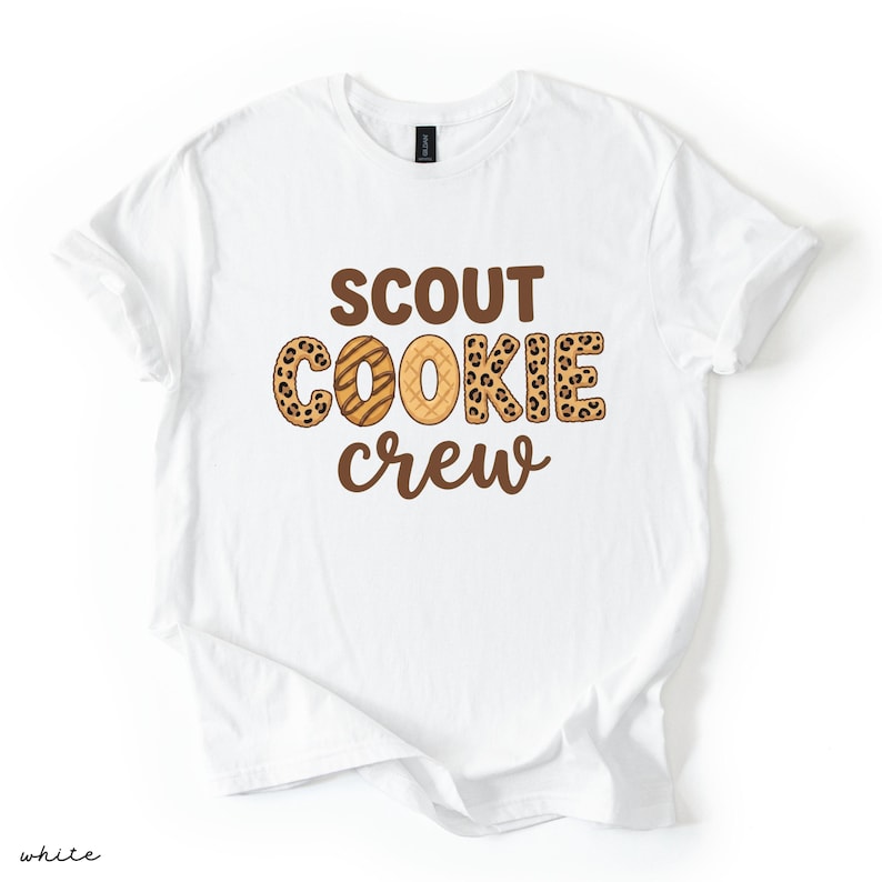 May include: White t-shirt with the words "SCOUT COOKIE crew" in brown lettering. The word "COOKIE" has a leopard print pattern and a cookie design. The shirt is made of a soft material and is perfect for cookie lovers.
