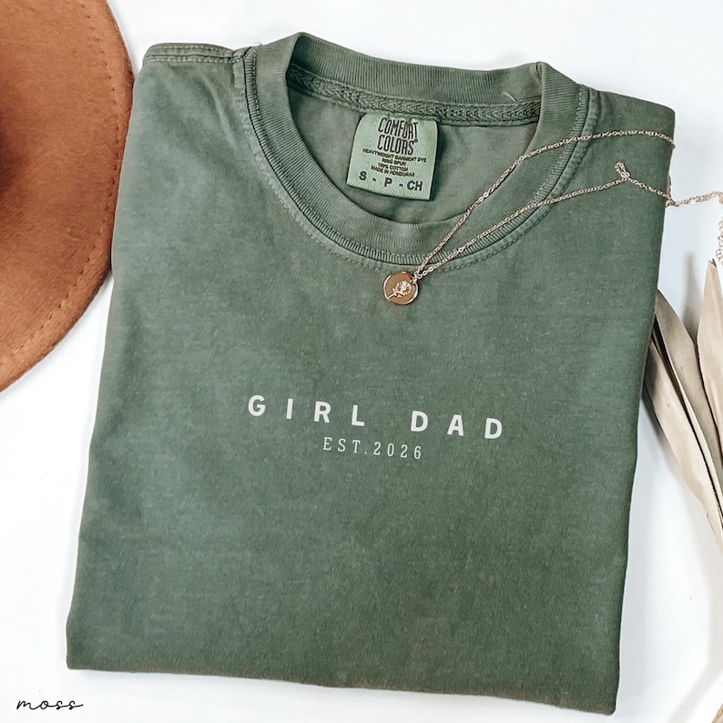 May include: An olive green t-shirt with the text "GIRL DAD EST. 2026" in white. A gold necklace with a pendant rests on the shirt. The shirt is folded and has a tag from Comfort Colors.