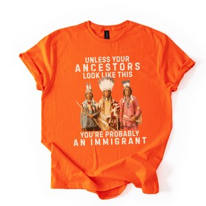 May include: Bright orange t-shirt with the text "UNLESS YOUR ANCESTORS LOOK LIKE THIS YOU'RE PROBABLY AN IMMIGRANT" in white. The shirt features a graphic of three Native American figures in traditional clothing.
