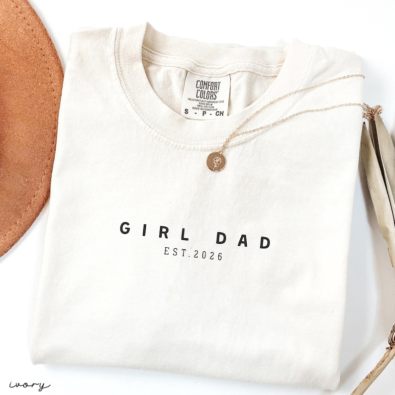 May include: An ivory-colored t-shirt with the words "GIRL DAD EST. 2026" printed in black. A gold-colored necklace with a circular pendant rests on the shirt. The shirt is folded and has a tag that says "COMFORT COLORS".