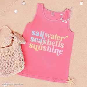 May include: A pink tank top with the words "saltwater seashells sunshine" in pastel colors. A woven bag and a seashell necklace are next to the tank top. The image is on a sandy surface with starfish.