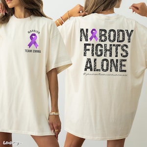 Pode incluir: T-shirt cor marfim com uma fita de conscientiza&ccedil;&atilde;o roxa e as palavras "WARRIOR" e "TEAM EMMA" na frente. A parte de tr&aacute;s da camisa apresenta o texto "NOBODY FIGHTS ALONE" com uma fita roxa e a hashtag "#pancreaticcancerawareness."