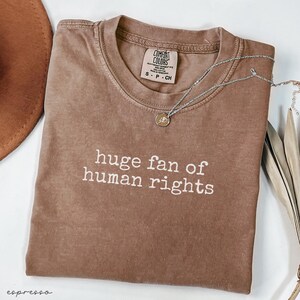 May include: Espresso-colored t-shirt with the text "huge fan of human rights" in white. The shirt has a crew neck and a Comfort Colors tag. A silver necklace with a small pendant is draped across the shirt.