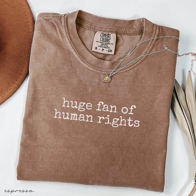 May include: Espresso-colored t-shirt with the text "huge fan of human rights" in white. The shirt has a crew neck and a Comfort Colors tag. A silver necklace with a small pendant is draped across the shirt.