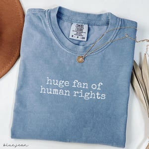 May include: A blue Comfort Colors t-shirt with the phrase "huge fan of human rights" in white lettering. A gold necklace with a pendant rests on the shirt. The shirt is folded and has a small tag.