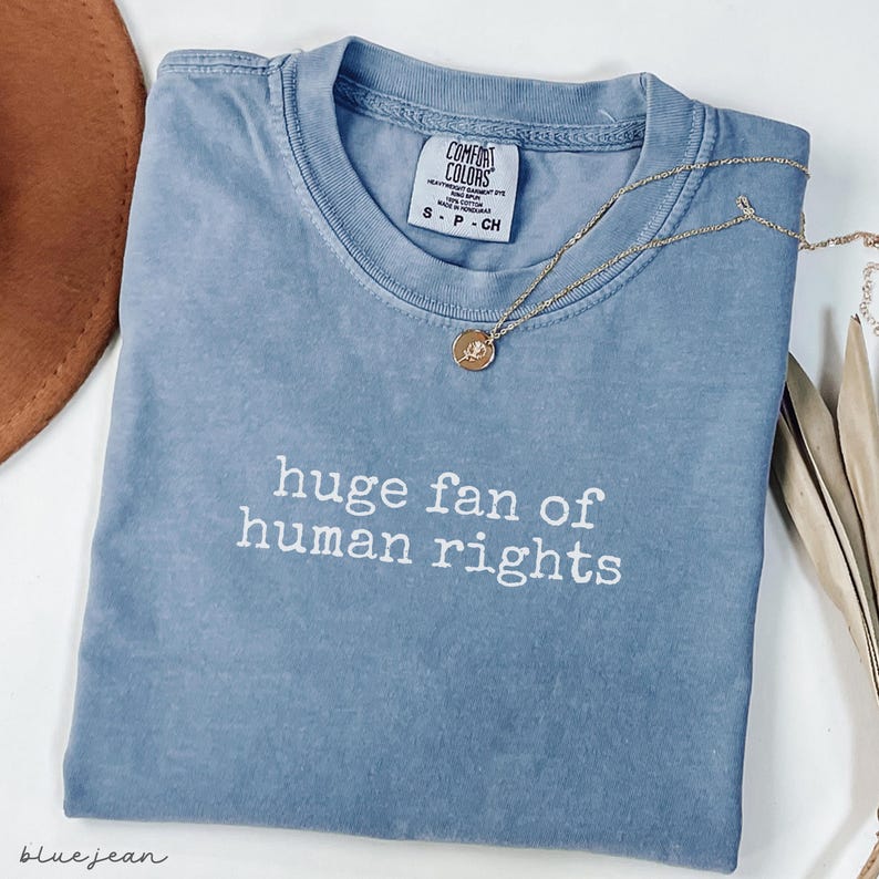 May include: A blue Comfort Colors t-shirt with the phrase "huge fan of human rights" in white lettering. A gold necklace with a pendant rests on the shirt. The shirt is folded and has a small tag.
