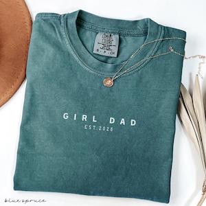 May include: A teal Comfort Colors t-shirt with the text "GIRL DAD EST. 2026" in white. A gold necklace with a pendant rests on the shirt. The shirt is folded on a white surface, with a brown hat and dried leaves in the background.