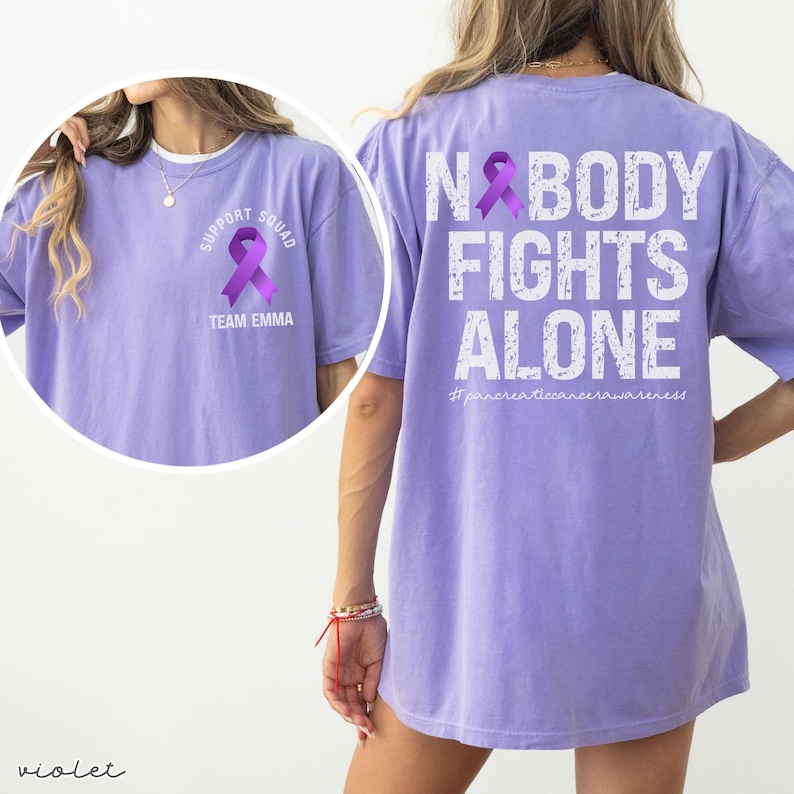 Pode incluir: Camiseta violeta com a frase branca &laquo;NOBODY FIGHTS ALONE&raquo; nas costas, junto com um la&ccedil;o roxo. A frente da camiseta apresenta um la&ccedil;o roxo e as palavras &laquo;SUPPORT SQUAD TEAM EMMA&raquo;. A hashtag #pancreaticcancerawareness tamb&eacute;m &eacute; vis&iacute;vel.